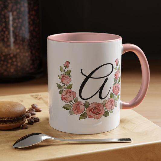 White mug with pink interior and handle, featuring floral design and letter 'A', on a wooden surface with coffee beans and a spoon.