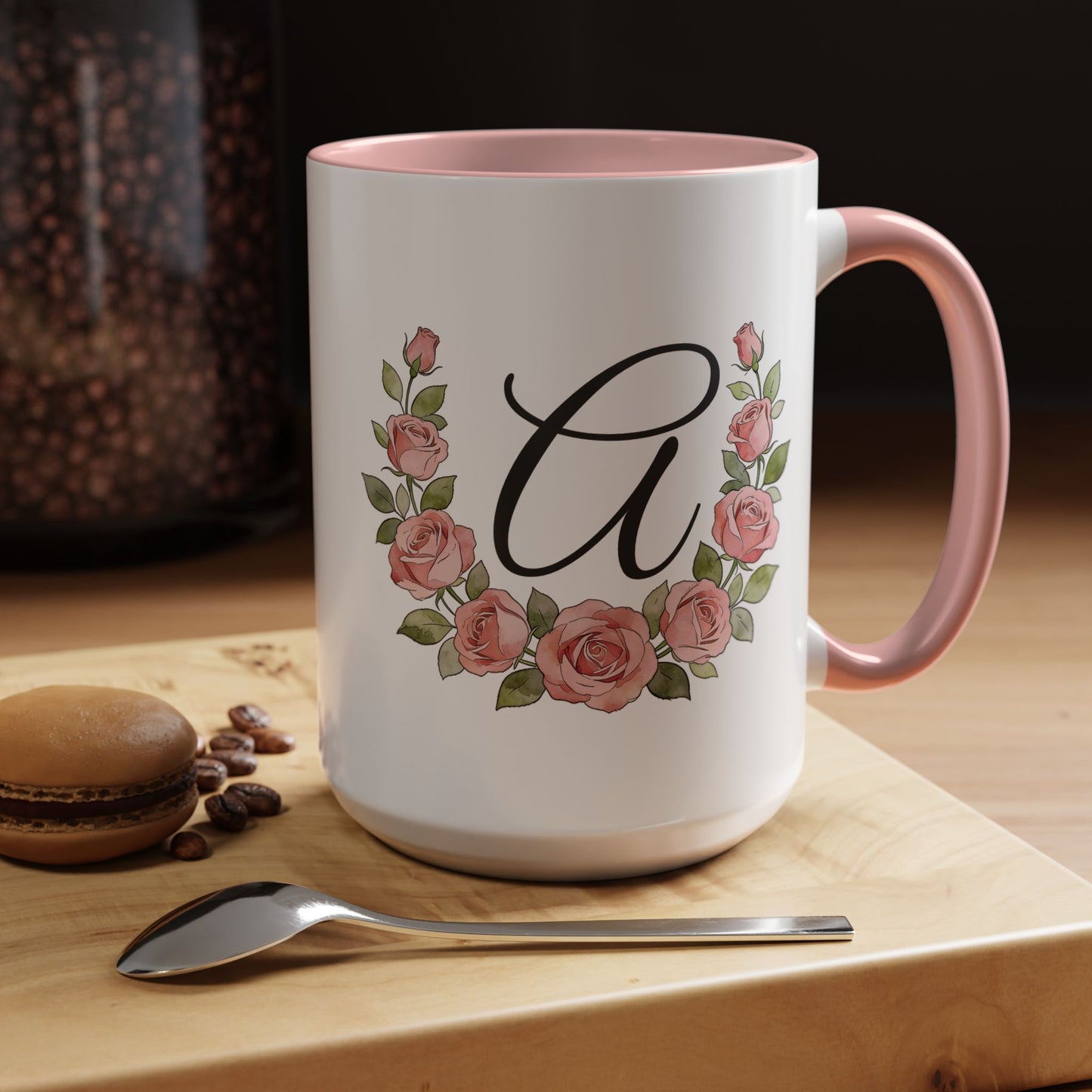 White mug with pink interior and handle, featuring a floral design and letter 'A', on a wooden surface with coffee beans and a macaron.